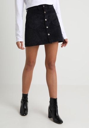 Zwarte corduroy minirok met voorzakken en knoopsluiting, gecombineerd met een wit overhemd met lange mouwen en glanzende zwarte enkellaarzen.