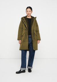Parka verde oliva con fodera in pelliccia, due tasche frontali, vita regolabile con cordoncino e cappuccio. Indossato sopra un top nero e jeans blu.