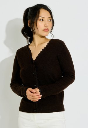 Mujer con cabello negro recogido en una coleta que lleva un cárdigan marrón oscuro abotonado y pantalones blancos, mirando hacia su izquierda con las manos entrelazadas.