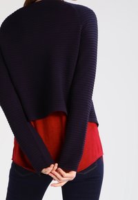 Námořnický žebrovaný svetr vrstvený přes červenou košili, s volným střihem a dlouhými rukávy. Lem svetru končí těsně nad pasem košile.