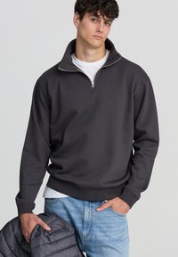 Felpa in carbone con colletto a mezza zip, maniche lunghe e polsini a coste; indossata sopra una t-shirt bianca, abbinata a jeans azzurri chiari.