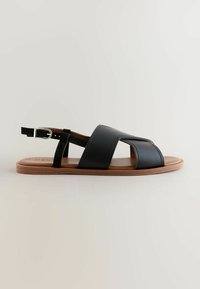 Sandalo in pelle nera con cinturini incrociati, fibbia regolabile alla caviglia e suola in gomma marrone. Design piatto per una facile calzata.