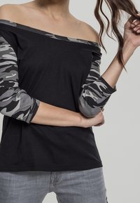 Kvinna med en svart off-shoulder top med grå kamouflagemönstrade ärmar och ljusgrå jeans, som håller en hårslinga med ena handen.