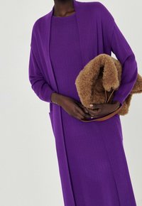 Modelka v fialových, žebrovaných šatech aodpovídajícím svetru drží chlupatou hnědou tašku s koženým popruhem před neutrálním pozadím.