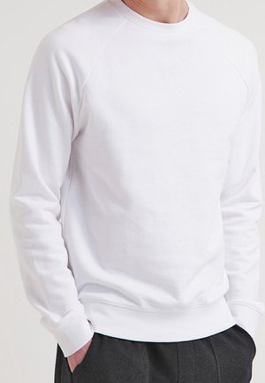 Mies, joka pukeutunut yksinkertaiseen valkoiseen pitkähihainen collegepaitaan ja tummiin housuihin, kädet taskuissa, kuvattu kaulasta lantiolle.