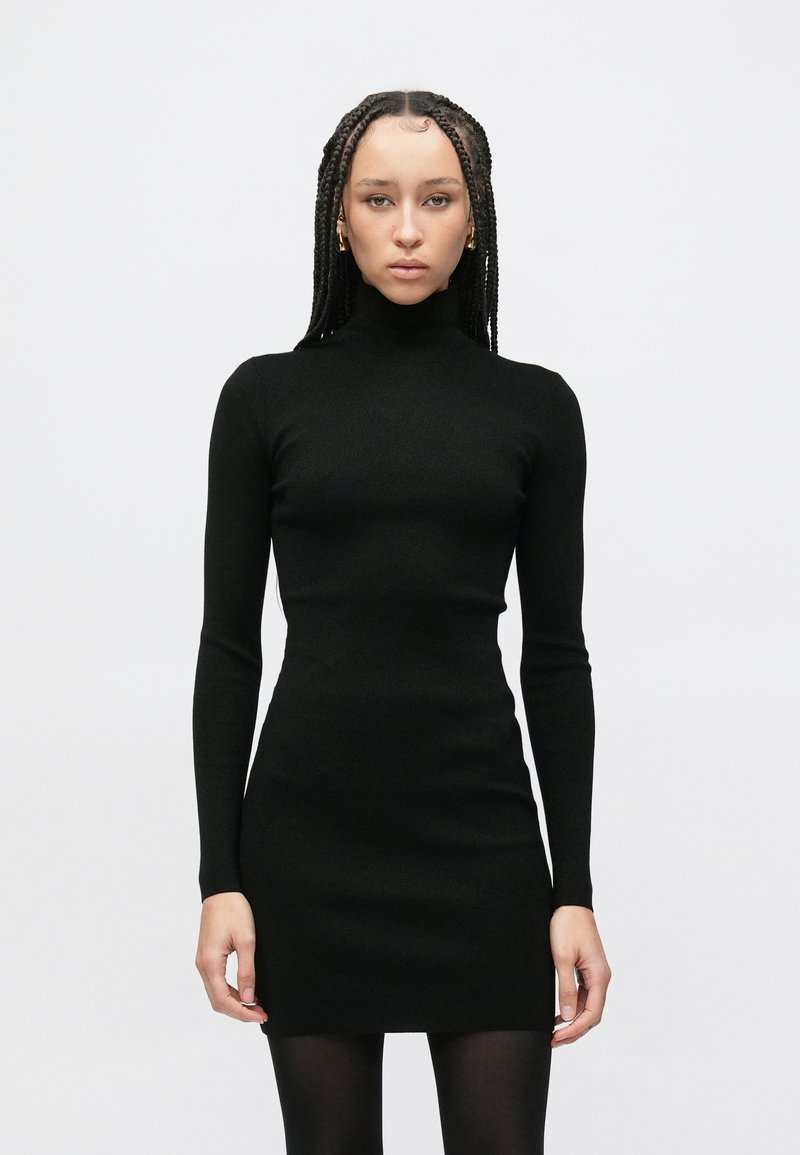 Czarna dopasowana sukienka z wysokim dekoltem, długimi rękawami i gładką fakturą. Noszona z czarnymi rajstopami, podkreślająca smukłą sylwetkę.