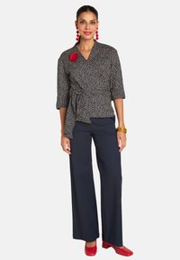 Schwarz-weiß gemusterte Wickelbluse mit Dreiviertelärmeln, kombiniert mit navyblauen weitgeschnittenen Hosen und roten Blockabsatzschuhen. Mit einem roten Blumenakzent.