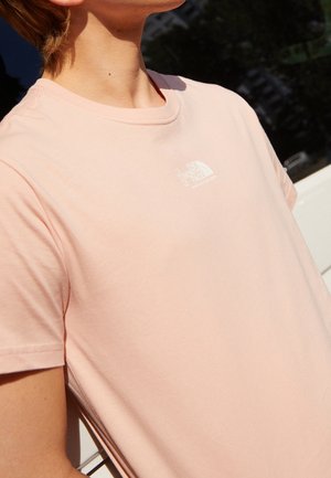 Light pink cotton t-shirt with short sleeves, crew neck, and a small white logo on the upper left side. Smooth texture, casual fit.
