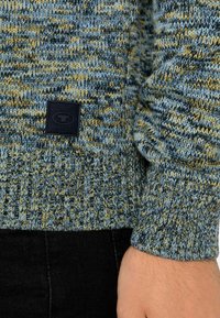 Maglione lavorato a maglia caratterizzato da una miscela di filati blu, giallo e grigio, con polsini a coste e un'etichetta nera del marchio cucita all'orlo.