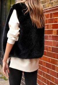Eine Person trägt eine schwarze Kunstpelz-Weste über einem weißen Sweatshirt und schwarzen Hosen und steht neben einer roten Backsteinmauer im Freien.