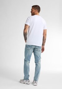 T-shirt bianco in cotone con maniche corte, abbinato a jeans di denim blu chiaro e sneakers sportive bianche. La vista posteriore mostra i tatuaggi sul braccio.