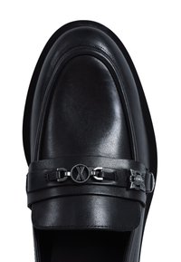 Mocasín de cuero negro con punta redonda, diseño elegante y una correa decorativa con acentos metálicos, que incluye un emblema circular y herrajes.