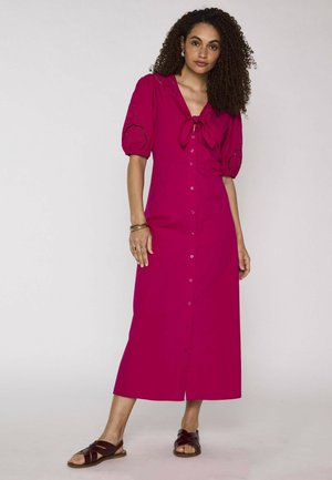 Femme aux cheveux bouclés portant une robe midi magenta boutonnée avec des manches bouffantes et des sandales noires, debout devant un fond uni.