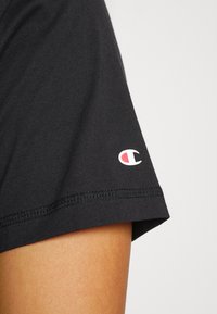 Zbliżenie na czarną koszulkę z krótkim rękawem na ramieniu osoby, z małym logo Champion w pobliżu dolnego brzegu.