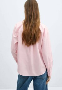 Rosa Button-up-Hemd aus Baumwolle mit langen Ärmeln und entspannter Passform, getragen über blauen Jeans. Einfaches Design, ohne sichtbare Muster.