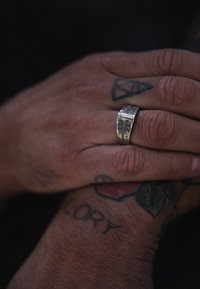 Sidabrinis žiedas su išgraviruotu delno medžio raštu, padėtas ant rankos su matomais tatuiruotiais. Odos tekstūra šiurkšti, rodo smulkius detalės.