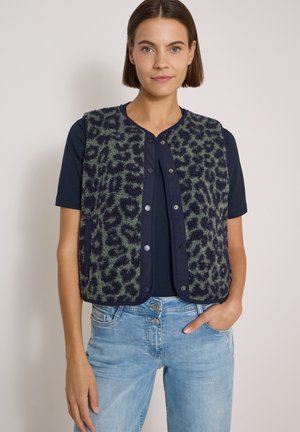 Kvinde med kort brunt hår iført en navyblå t-shirt, blå leopardmønstret ærmeløs vest og lyseblå jeans, stående mod en ensfarvet baggrund.