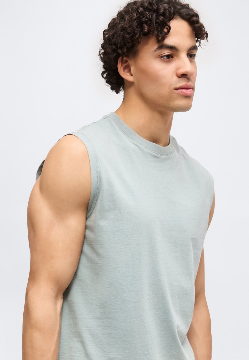Joven con cabello rizado que lleva una camiseta sin mangas de color gris claro, mirando ligeramente hacia la derecha contra un fondo liso.