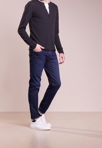 Svart långärmad henley-tröja med tre knappar, i kombination med mörkblå slim-fit-jeans och vita sneakers, mot en neutral bakgrund.
