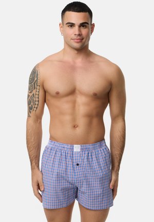 Boxers à carreaux bleus et rouges en tissu doux, avec une taille élastique et un détail de bouton à l'avant. Modèle présenté avec une expression neutre.