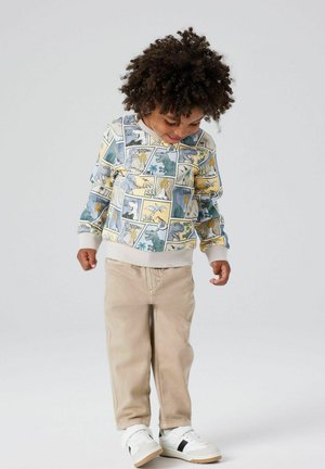 Hettegenser med tegneseriestil trykk i gule, blå og kremfargede toner, kombinert med beige bukser og hvite joggesko med svarte detaljer.