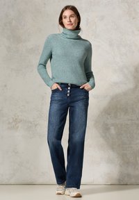 Türkiser Strickpullover mit Rollkragen und geripptem Muster, kombiniert mit hoch sitzenden blauen Denim-Weiter-Hosen mit Knopfleiste und seitlichen Taschen.