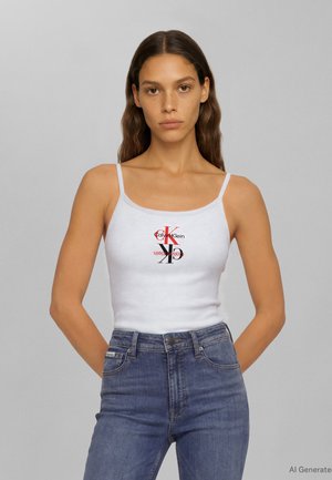 Frau mit langem, welligem Haar, die ein weißes ärmelloses Calvin Klein Top und hoch taillierte blaue Jeans trägt, steht vor einem schlichten grauen Hintergrund.