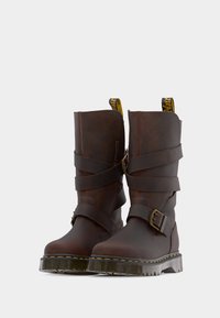 Ζευγάρι από σκούρα καφέ δερμάτινες μπότες μέσης μοσχαριού με διπλές λουρίδες με αγκράφες και παχιά ημιδιάφανα πέλματα με λουρίδες, εμφανιζόμενες σε ανοιχτό γκρι φόντο.