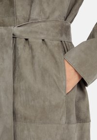 Suede trench coat in muted gray with a textured finish. Features a tied belt, side pocket, and visible stitching detail along the seams.