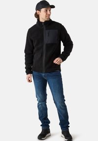 Svart fleecejacka med hög krage, dragkedjeficka på bröstet och två sidofickor, tillsammans med blå jeans och mörka sportskor.