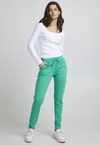 Rohelised spordipüksid elastse vöökoha ja nööriga, koos valge ribakujulise pika varrukaga tipuga. Mudel seisab neutraalses taustas.