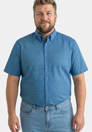Uomo con capelli biondi corti e barba indossa una camicia blu a maniche corte con fantasia e jeans azzurri, in piedi davanti a uno sfondo uniforme.