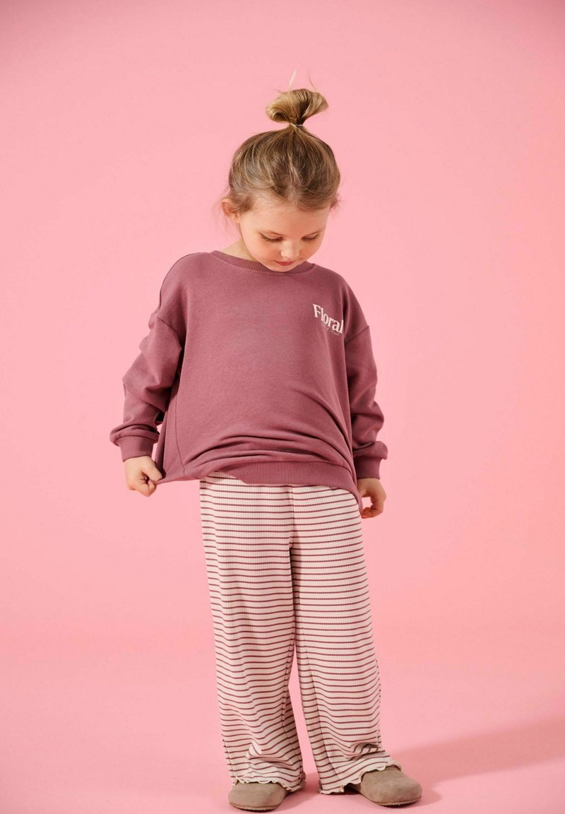 Νεαρή κοπέλα με σινιόν κότσο, φοράει φαρδιά μωβ μπλούζα και ριγέ φαρδιά παντελόνια, στέκεται μπροστά σε ροζ φόντο, κοιτάζοντας προς τα κάτω.