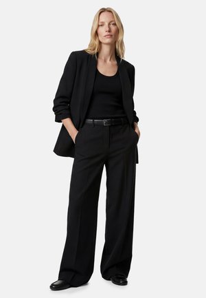 Blondynka stojąca z rękami w kieszeniach, ubrana w czarną marynarkę, czarny top, szerokie czarne spodnie, czarny pasek oraz czarne buty.