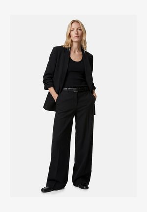 Blonde Frau steht mit den Händen in den Taschen, trägt einen schwarzen Blazer, ein schwarzes Tanktop, weite schwarze Hosen, einen schwarzen Gürtel und schwarze Schuhe.