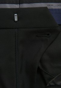 Schwarzes Stoffkleidungsdetail mit einem Reißverschluss, metallischen Beschlägen und einem kleinen, in Schwarz gestickten Logo. Texturierte Bereiche in Grau und Marineblau.