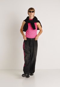 Pinkes Tanktop, schwarze übergroße Jeans mit pinkem Grafikdruck, schwarzer Pullover locker über den Schultern getragen, Sonnenbrille und schwarze Schuhe.