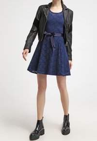 Robe en dentelle navy avec jupe évasée, associée à une veste de moto en cuir noir et des bottes noires à talons épais. Texture douce, taille cintrée.