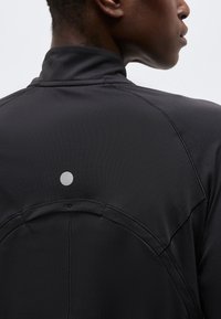 Κοντινό πλάνο ενός ατόμου που φοράει μαύρο μπουφάν με ψηλό γιακά, με ορατή ραφή και έναν μικρό ανακλαστικό κύκλο στο πάνω μέρος της πλάτης.