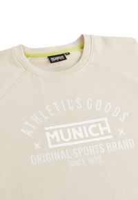 Sweat-shirt beige avec un col rond et des manches raglan. Présente un branding en texte blanc : "ATHLETICS GOODS MUNICH ORIGINAL SPORTS BRAND."