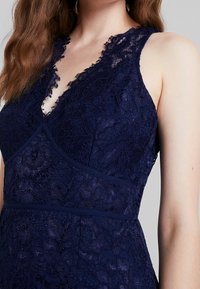 Vestido de encaje en azul marino con escote en V profundo, patrones florales y un diseño ajustado con ribetes contrastantes alrededor del busto y la cintura.