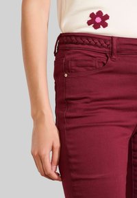 Primo piano di una mano di una persona appoggiata accanto a pantaloni borgogna con una cintura intrecciata, indossando un top crema con un ricamo di fiori rossi e rosa.