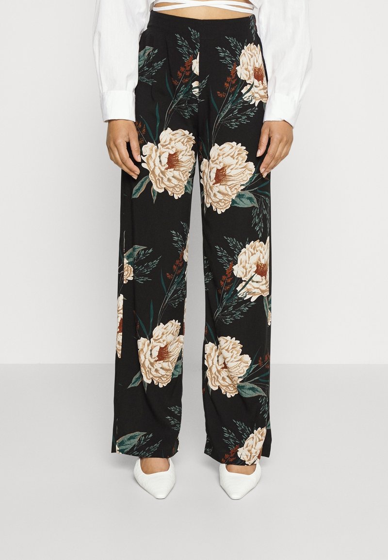 Femme portant un pantalon noir à jambes larges avec un grand imprimé floral crème, associé à des chaussures blanches à bout pointu et un top court blanc.