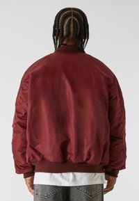 Burgunderfarvet bomberjakke med en glat, skinnende overflade, ribbestrikkede manchetter og waistband. Bagfra ses et minimalistisk design uden synlig branding.