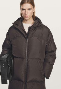 Giovane che indossa un lungo giaccone puffer nero con cappuccio, mentre porta una borsa in pelle nera, in piedi davanti a uno sfondo chiaro e semplice.