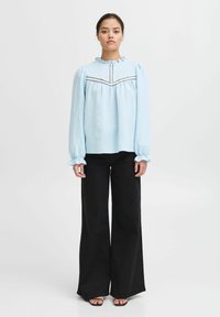 Blusa a maniche lunghe azzurro chiaro con colletto arricciato e placket. Presenta cuciture nere a contrasto e una vestibilità comoda. Abbinata a pantaloni a gamba larga neri.