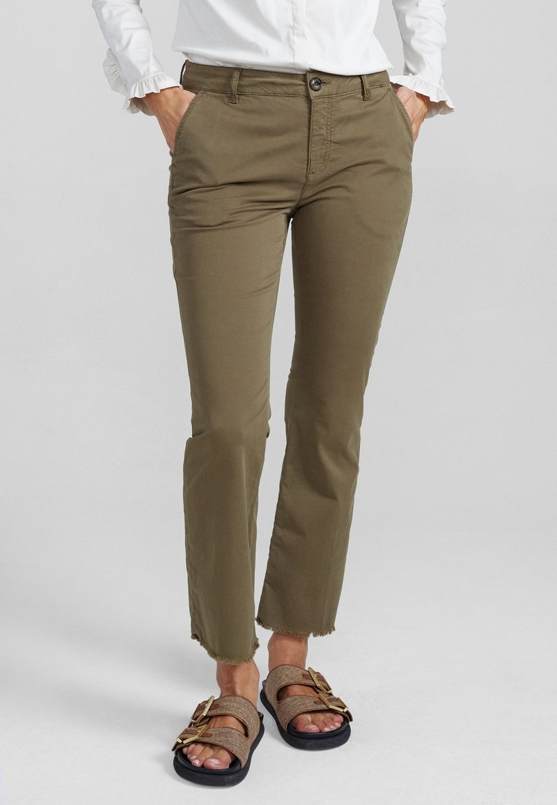 Personne portant un pantalon court vert olive aux ourlets effilochés, une chemise blanche à manches longues, et des sandales brunes à double boucle. Mains dans les poches.