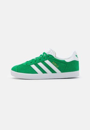 Baskets Adidas Gazelle vertes avec des bandes blanches, semelle blanche et languette au talon sur fond blanc uni, vue de côté.
