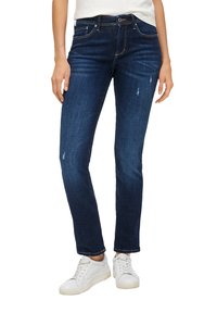 Jeans en denim bleu foncé avec une coupe slim, présentant des effets de frottement subtils et des détails usés. Portés avec des sneakers blanches.
