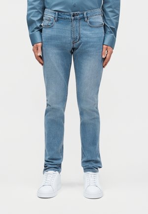 Άνδρας που φοράει στενά τζιν σε ανοιχτό γαλάζιο χρώμα, μπλε πουκάμισο με κουμπιά και λευκά αθλητικά παπούτσια, στέκεται μπροστά σε ένα απλό φόντο.